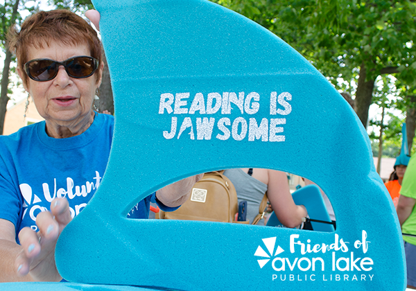 a volunteer member is holding a shark fin hate that says "reading is jawsome". The Friends of Avon Lake Public Library" is displayed o the bottom.